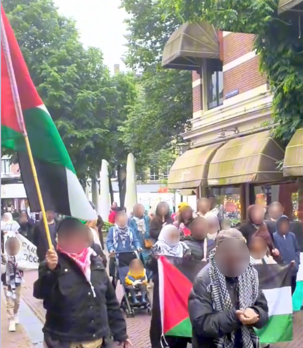 Tientallen mensen lopen op een heldere dag op straat in een binnenstad met Palestijnse vlaggen, spandoeken, en kuffiya om.