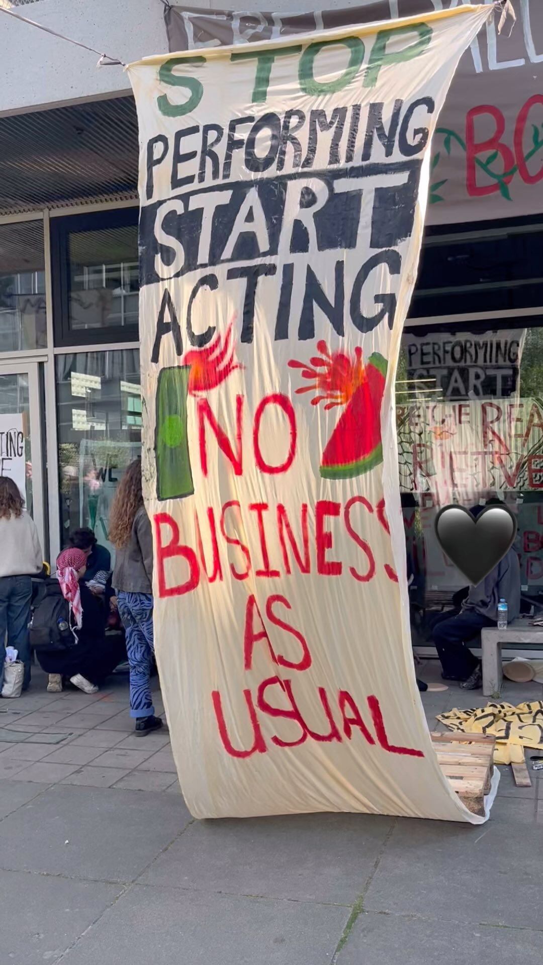 Een gebouw met grote ramen en op de gevel verschillende kleurrijke spandoeken en verschillende mensen voor het gebouw landslopen of zitten. Central in beeld is een lang spandoek met daarop in het Engels in groene, zwarte en rode letters de tekst: 'Stop met optreden, begin met handelen. Doe niet alsof er niks aan de hand is', plus en watermeloen en nooduitgang met vuur als illustraties.