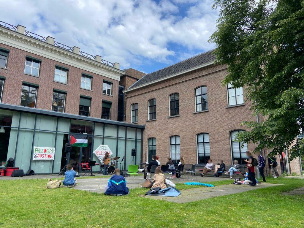 Op een binnenplaats met plaveisel, gras en bomen, voor een bakstenen en glazen gebouw met hoge ramen, staat of zit een tiental mensen. De mensen richten zich naar het midden, waar een persoon met een gitaar achter een microfoon zingt of spreekt. Rondom hen staan ook een drumstel, toetsenbord en luidsprekers. Achter hen hangt een Palestijnse vlag en spandoeken met de teksten (in het Engels): 'Vrijheid en gerechtigheid' en 'Bevrijd Palestina'. De lucht is halfbewolkt.