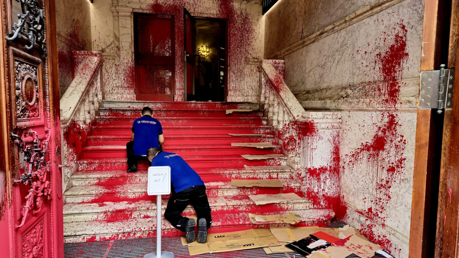 De klassiek marmeren ingang van een gebouw is met heldere rode verf beklad. Op de traptreden liggen stukken karton, waar mensen op kunnen lopen zonder in de verf te trappen. Twee personen in een blauw shirt en zwarte broek zijn bezig met de schoonmaak. Een bord bij de ingang draagt, in het Engels, de boodschap 'Pas op!'.