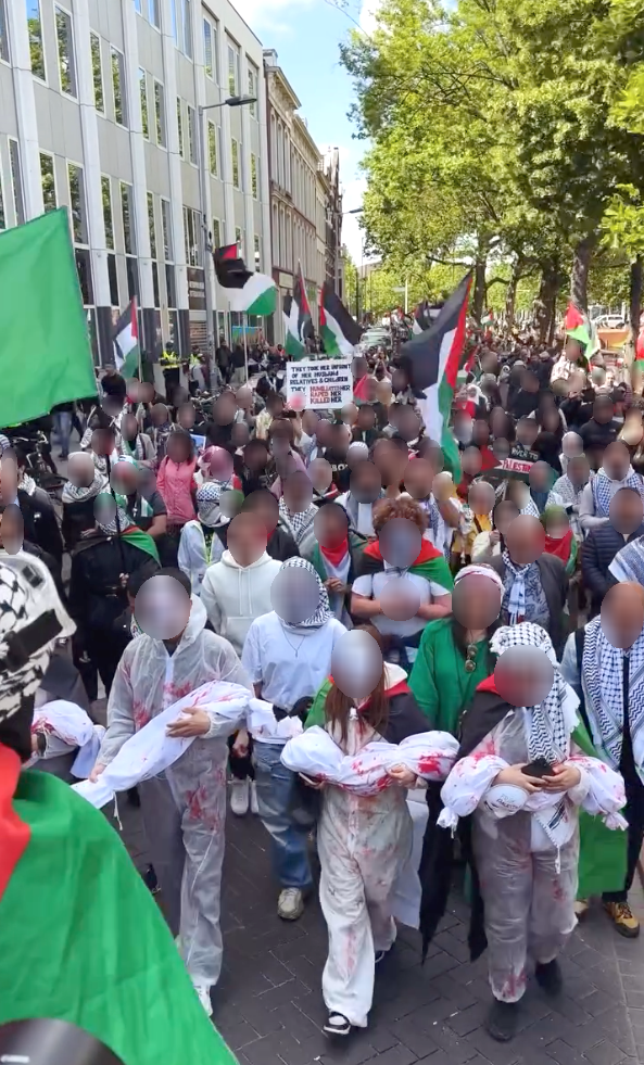 Honderden mensen lopen op een zonnige dag op straat langs een appartementgebouw en bomen. Zij zwaaien met Palestijnse vlaggen en dragen borden. Op de voorgrond hebben drie mensen beelden in hun armen van bebloede baby's in lijkzakken.