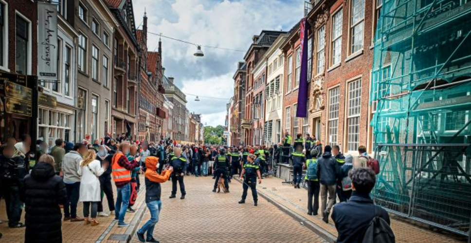 Een grote menigte mensen verzamelt zich midden op straat en langs de trottoirs van een stadsstraat. De straat is geplaveid met lichtbruin-oranje stoeptegels, met aan beide kanten van de straat grote bakstenen gebouwen met meerdere verdiepingen. Eén gebouw aan de rechterkant van de straat is deels bedekt met steigers en een blauwgroen vangnet. Een aantal mensen draagt borden en spandoeken. Vanaf een bovenverdieping van een gebouw aan de rechterzijde van de straat hangt een lang, gedrapeerd spandoek aan de gevel. Er is heel veel politie aanwezig, die zich verzamelt rond de ingang van het gebouw met het spandoek.
