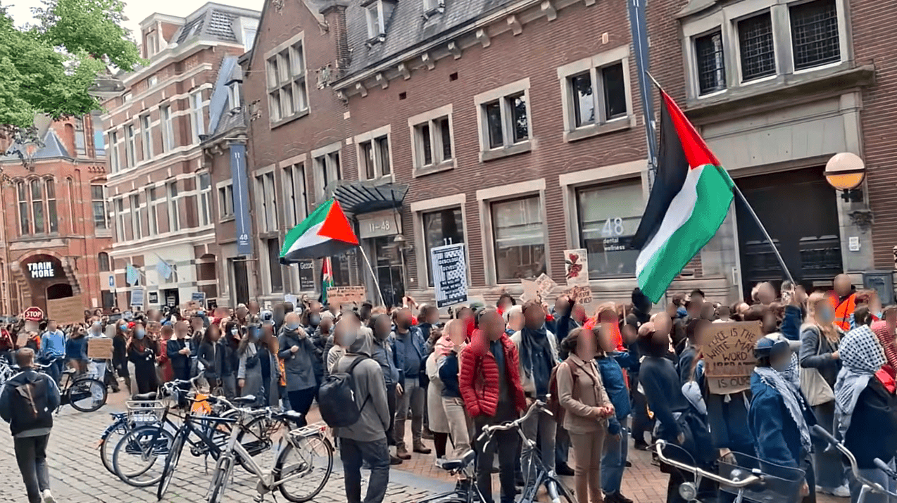 Honderden mensen staan op een grauwe dag in een oud stadscentrum met Palestijnse vlaggen en borden en spandoeken voor Palestina.