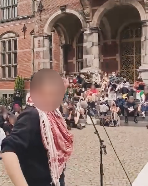 Een persoon staat voor een microfoon en kijkt over diens schouder achterom recht in de camera. Op de achtergrond zitten op een kort, oplopende trap zo rond de veertig mensen met Palestijnse vlaggen.