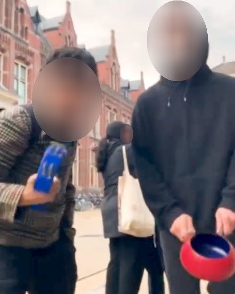 Twee mensen buigen zich op straat naar beneden richting de camera. De persoon links bespeelt een tamboerijn, en de persoon rechts bespeelt een pan.