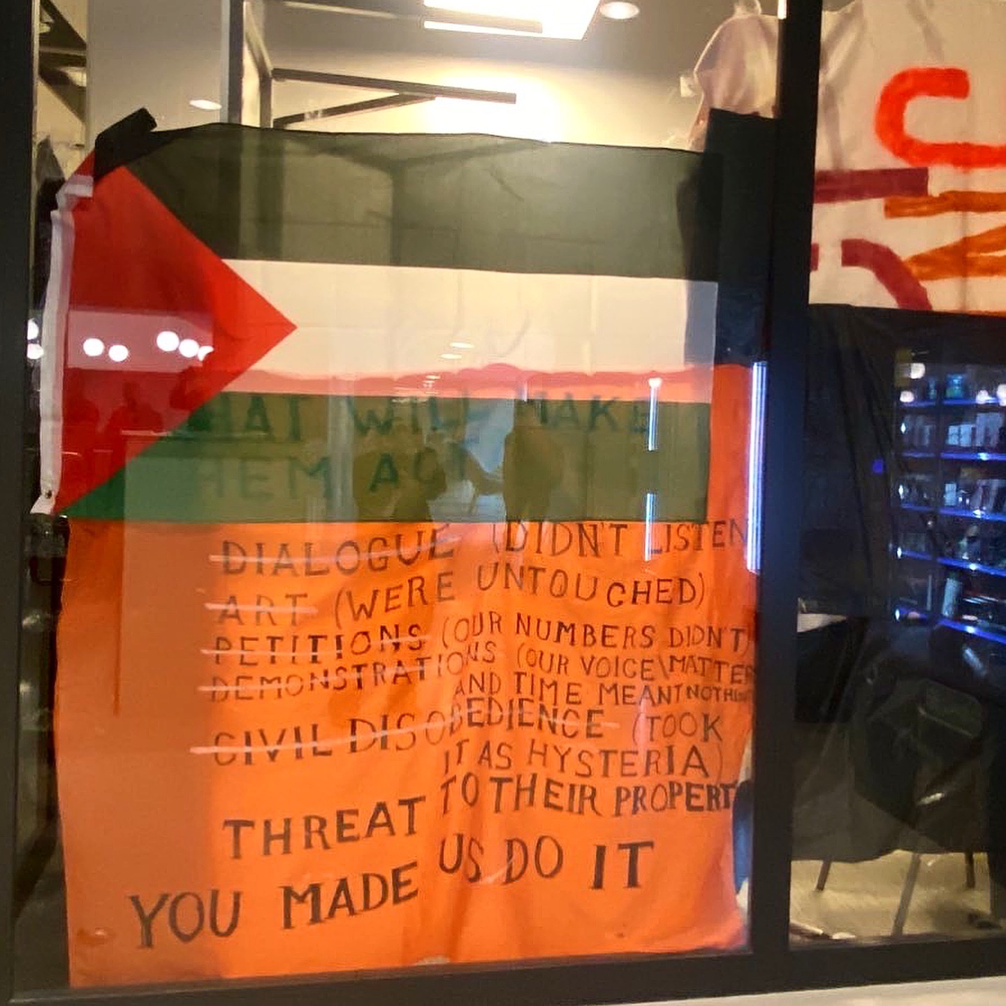 De glazen gevel van een ruimte met daarin stoelen, en kast met objecten, en felle plafondverlichting. Tegen het raam hangt een Palestijnse vlag en een oranje spandoek met daarop de tekst (in het Engels): 'Wat hen tot handelen zal aanzetten: dialoog (luisterden niet), kunst (werden er niet door geraakt), petities (onze aantallen maakten geen verschil), demonstraties (onze stem en tijd betekenden niets voor hen), burgerlijke ongehoorzaamheid (interpreteerden ze als hysterie), bedreiging van hun eigendom. Jullie hebben ons hiertoe aangezet.'