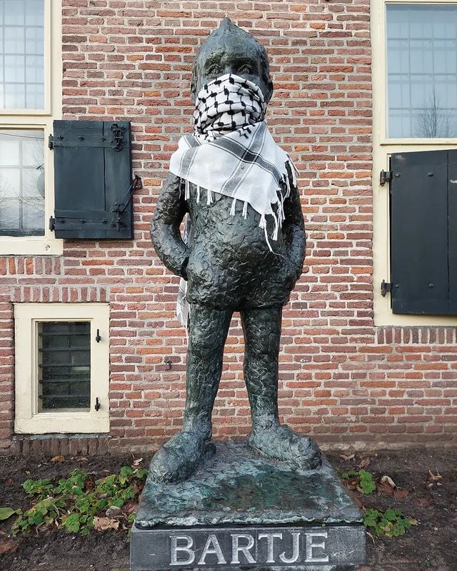Bronzen standbeeld van een kind op klompen, met een kort jasje aan en de handen in de zakken. De figuur heeft kort haar en kijkt recht vooruit. Over diens mond, neus en nek draagt het kind een wit-zwarte, stoffen kuffiya. Het beeld staat op een kleine verhoging in een plantsoen met voornamelijk aarde en enkele groen planten. Het beeld staat vlak voor een gebouw met rode bakstenen, en ramen met witte houten kozijnen en donkere luifels.