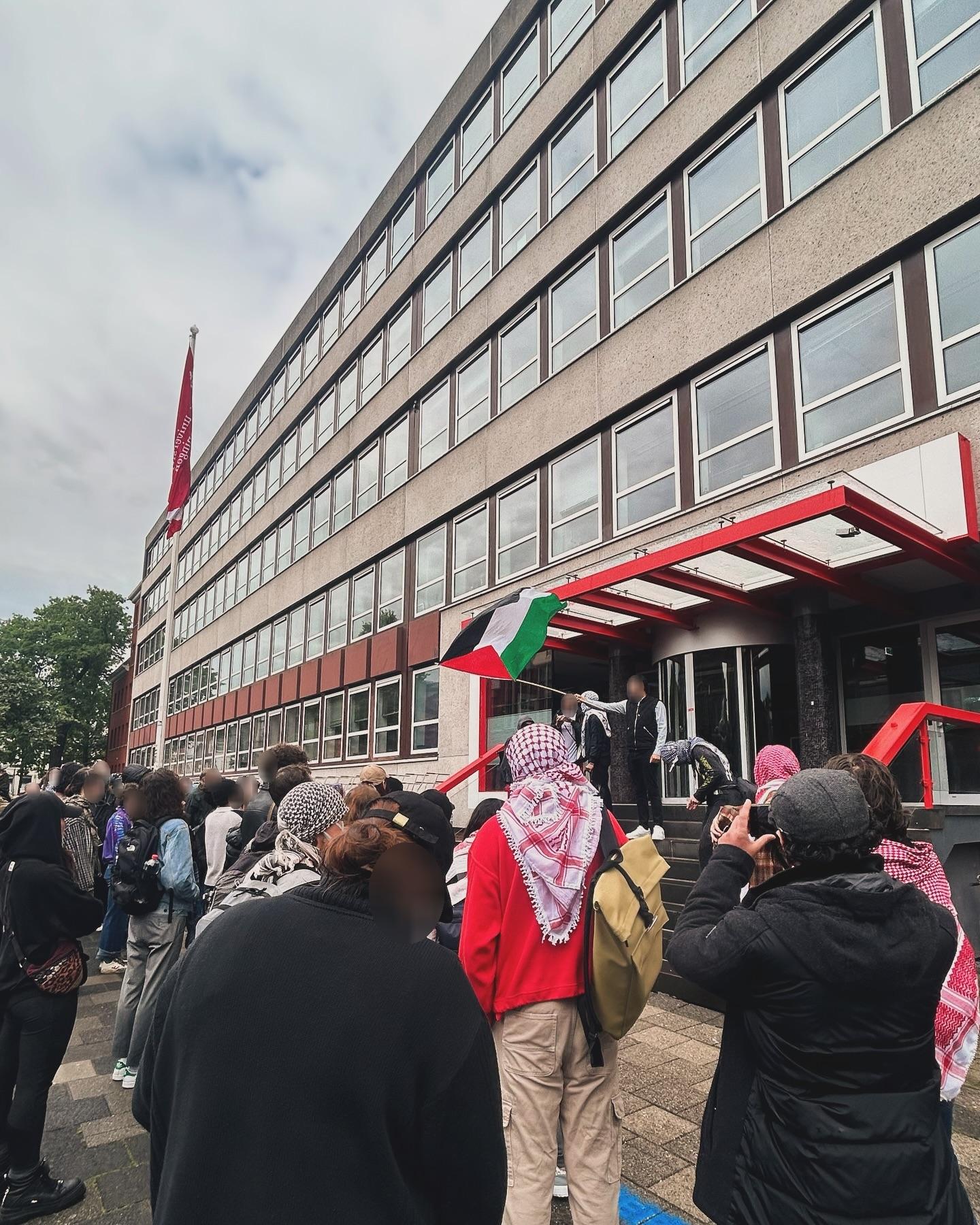 Tientallen mensen staan voor de ingang van een strak gebouw met meerdere verdiepingen. Verschillenden van hen dragen kuffiya. Op een verhoging bij de ingang van het gebouw zwaait een persoon met een Palestijnse vlag.
