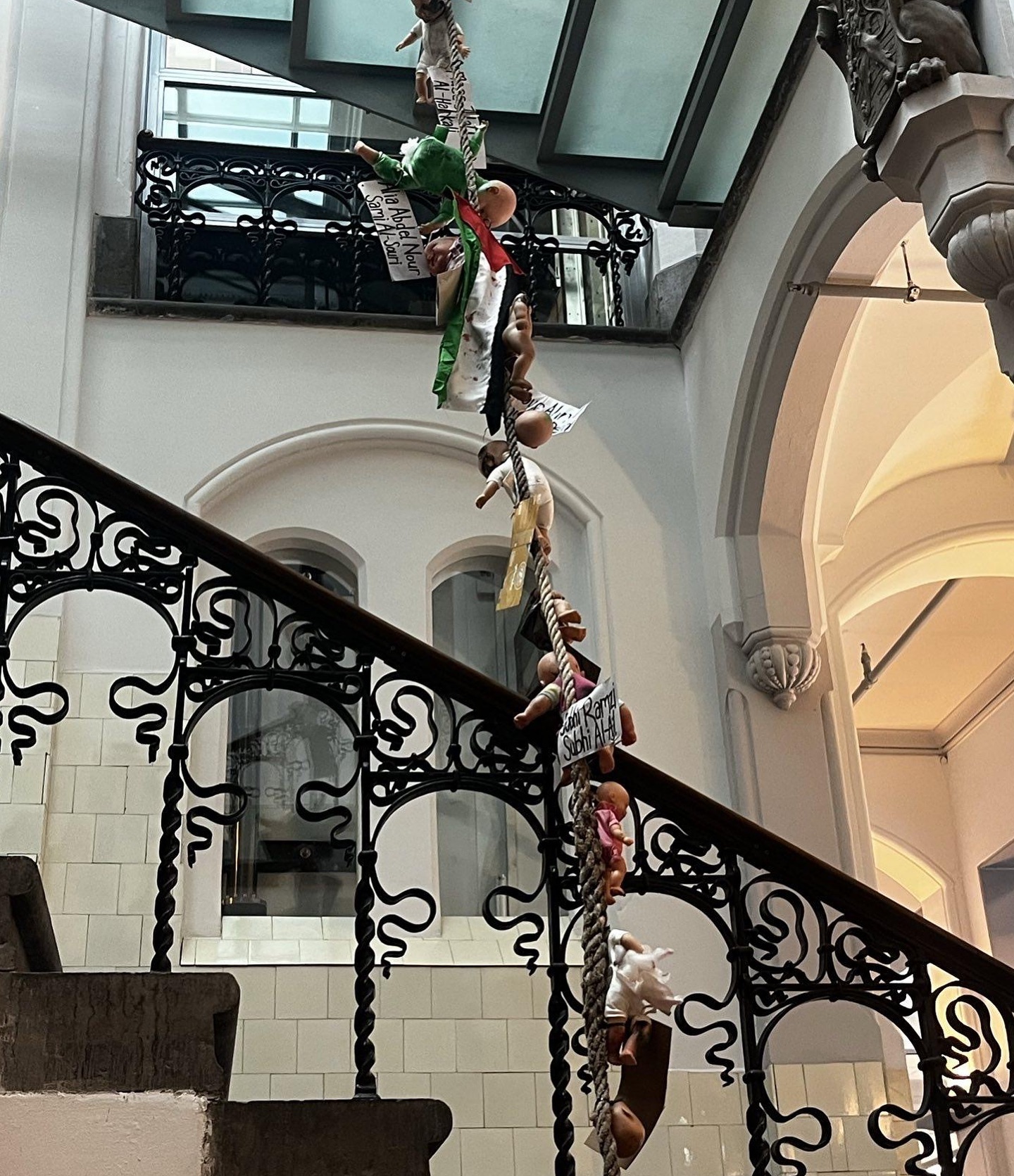 In een licht, klassiek trappenhuis met een trapleuning met metalen art-deco-elementen, hangt vanaf boven een touw met daaraan vastgeknoopt kinderpoppen en hier en daar een Palestijnse vlag. De poppen dragen briefjes met daarop namen, waaronder 'Alia Abdel Nour Sami al-Souri'.
