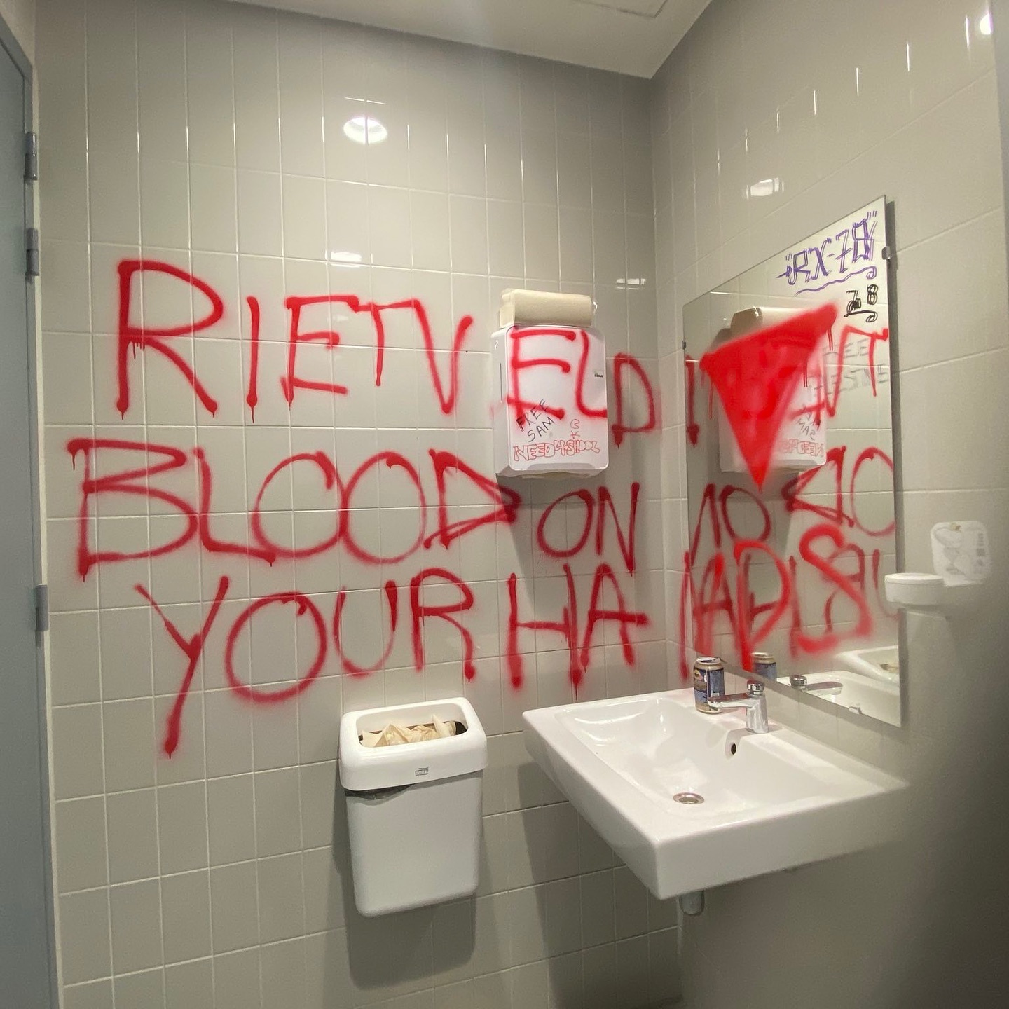 De hoek van een lichtgrijs betegeld toilet met wastafel, spiegel, handendroger en prullenbak. Strekkend over de hoek van de ruimte is met rode spuitverf in het Engels geschreven: 'Rietveld bloed aan je handen', met een rode, naar beneden gerichtte driehoek.