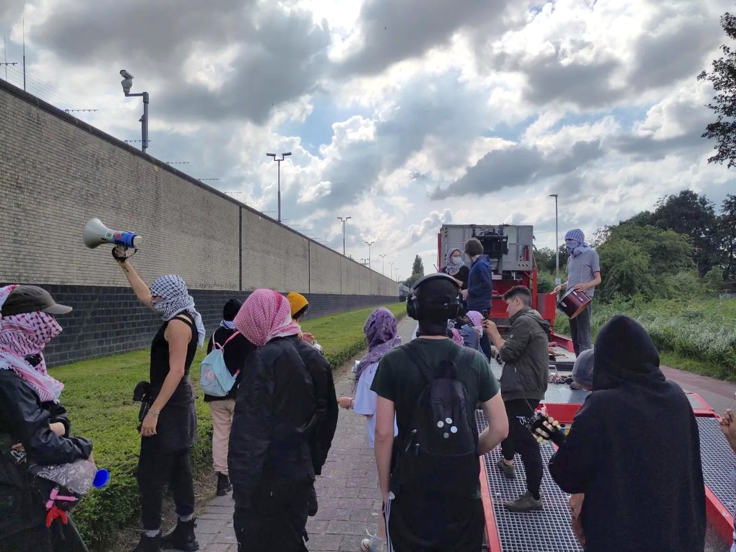 Een groep mensen met kuffiya om staan op een bewolkte dag op en rondom een kar langs een grasveldje buiten een gevangenismuur. Met een megafoon roepen ze richting de gevangenis. Ook bespelen mensen trommels, gitaren en andere instrumenten.