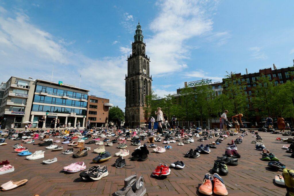 Op een groot, open plein, met aan de linkerkant een bankgebouw en aan de rechterkant een klokkentoren en commerciële gebouwen, staan op een zonnige, halfbewolkte dag duizenden paren schoenen uitgestald. Het hele plein is met precieze rijen van schoenparen bedekt. Verschillende mensen loopt tussen de schoenenrijen door, ofwel om ze te bekijken ofwel om ze neer te leggen.