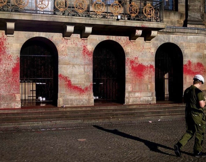 Een deel van een groot historisch gebouw met op de eerste verdieping een rijk-uitziend balkon bekleed met goudkleurige decoratie. Op de begane grond, tussen lichtbeige stenen muren, drie grote boogingangen met metalen hekken ervoor. De muren op de begane grond zijn rijkelijk besmeurd met bloedrode verf. Iets van het gebouw af loopt op de kleine kassieren een persoon in olijfgroene, militaire kleding met een witte helm op het beeld uit.