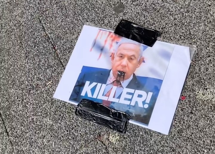 Bovenaanzicht op een afbeelding van massamoordenaar Benjamin Netanyahu waarbij zwarte olie uit diens mond komt en bloed op de achtergrond en op diens kleding zit is met zwart ductape op de stoep geplakt.