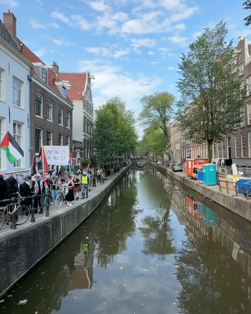 Zicht op een gracht met aan weerszijden panden en bomen. Het is een zonnige dag. Op de rechterkade staat voornamelijk bouwmateriaal. Over een brug in de verte en op de linkerkade marcheren duizenden mensen met Palestijnse vlaggen. Eén spandoek roept op (in het Engels): 'UvA, stop de leugens, verbeek alle banden'.