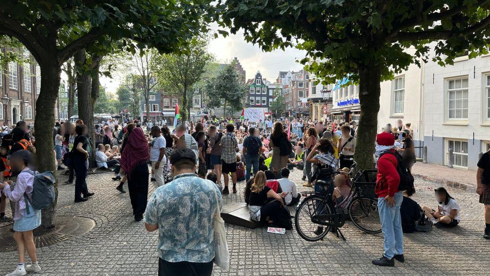 Een plein met verschillende bomen en kinderkopjes straatstenen, met links hoge bakstenen gebouwen en rechts witte stenen gevels. Op het plein staan en zitten honderden mensen, velen met borden, keffiyeh om en Palestijnse vlaggen. Op één spandoek staat in het Engels: 'UvA stop de leugens, verbreek alle banden'.