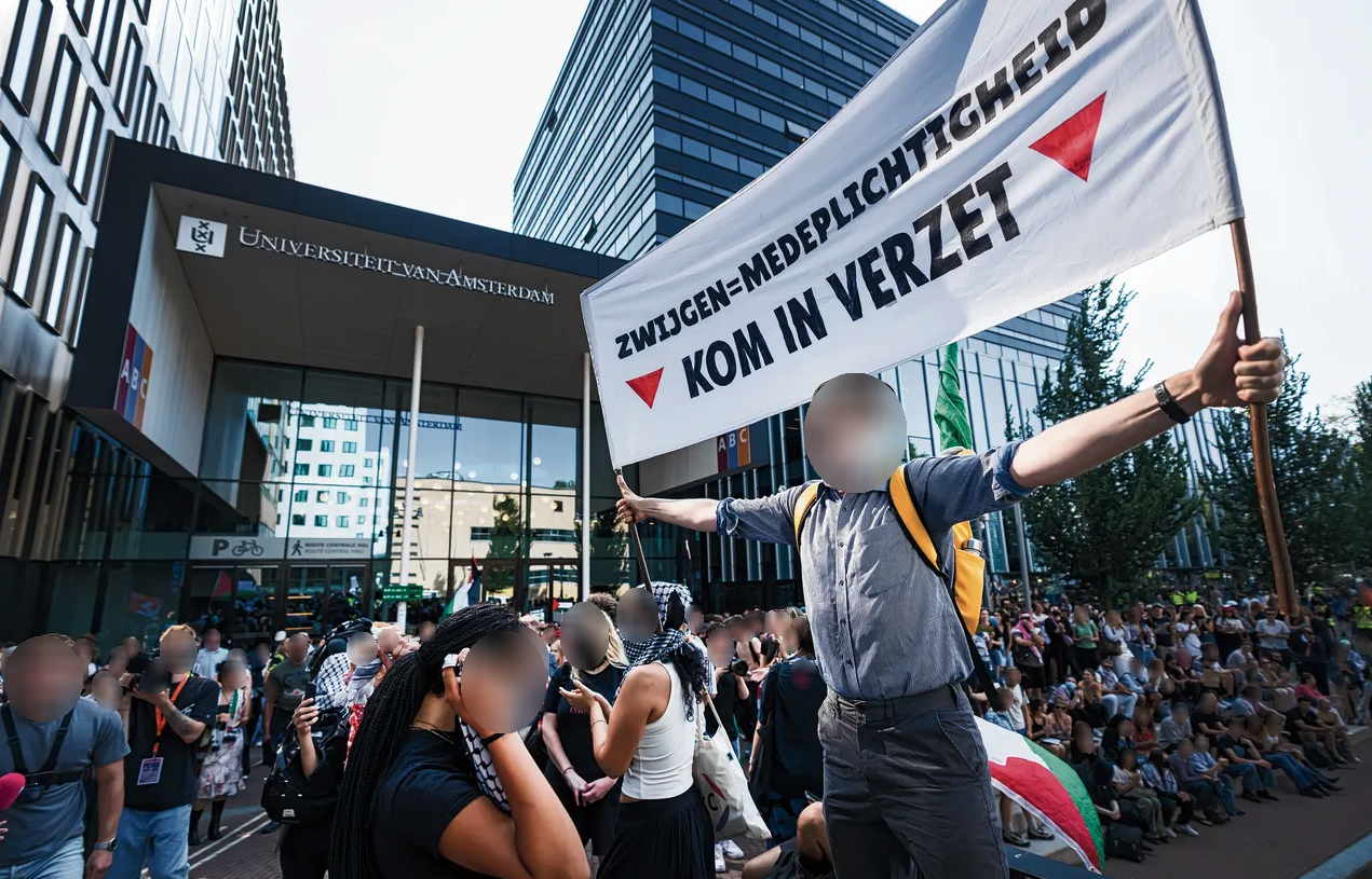Duizenden mensen staan en zitten op de stoep en kade voor een gebouw van met een reflecterende gevel met woorden 'Universiteit van Amsterdam'. Vele dragen Palestijnse vlaggen en keffiyeh. Vooraan in beeld staat een persoon met een spandoek met uitgestrekte armen, met daarop de tekst: 'Zwijgen is medeplichtigheid, kom in verzet', met rode, naar beneden gerichte driehoeken aan weerszijden van de tekst.