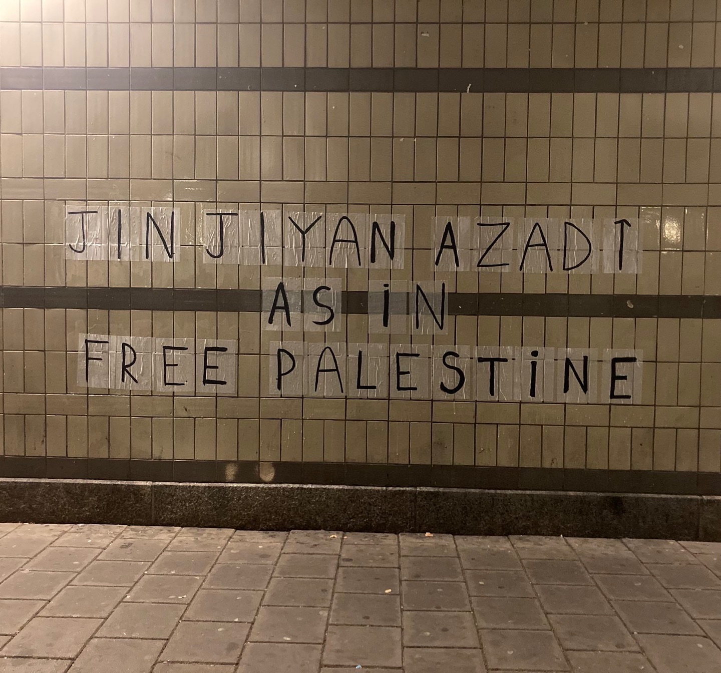 Een betegelde muur bij nachtverlichting. Op de puur zitten biljetten aangeplakt met verschillende losse letters, die zamen in het Koerdisch en Engels de zin vormen: 'Vrouw, leven, vrijheid, als in vrij Palestina'.