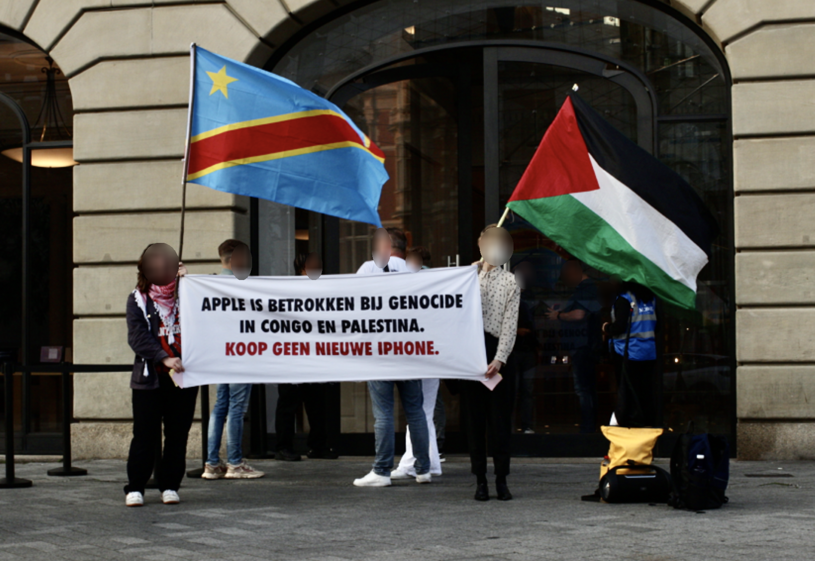 Twee mensen staan buiten op de stoep voor de hoge, glazen deuren van de ingang van een gebouw. Achter hen staat een bewaker met een blauw hesje aan. Eén van de personen heeft een Congolese vlag vast, de ander de Palestijnse. Samen dragen de personen een spandoek met daarop de in zwarte en rode letters de tekst: 'Apple is betrokken bij genocide in Congo en Palestina. Koop geen nieuwe iPhone.'
