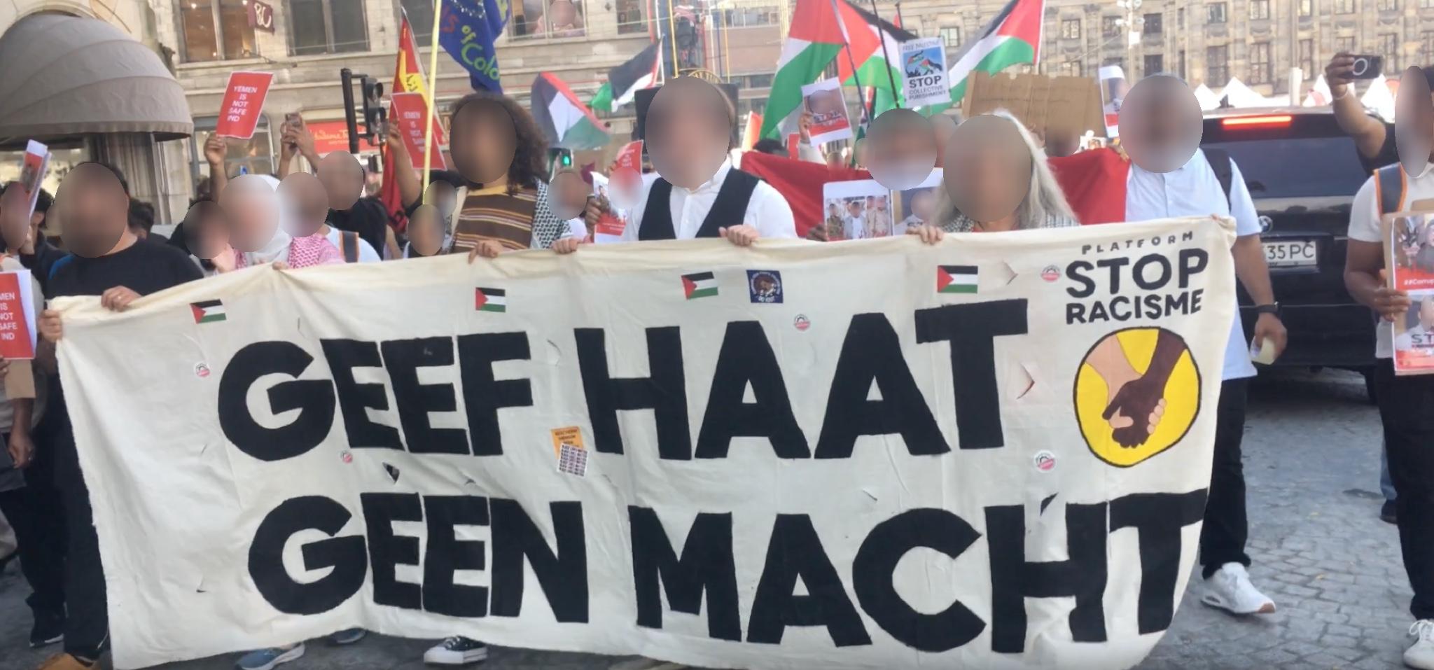 Een menigte van mensen loopt op straat in een stad op een lichte, zonnige dag, met auto's die langs hen heen gaan. Aan de voorkant lopen vijf mensen die samen een banner dragen. Op de banner staat de tekst 'Geef haat geen macht', met in de rechterbovenhoek van het spandoek een schildering van twee handen die elkaar schudden met de tekst 'Platform stop racisme' en daarnaast vele stickers met een Palestijnse vlag. De mensen die het spandoek dragen zingen of schreeuwen. In de menigte achter hen houden mensen borden, spandoeken, en Palestijnse vlaggen op.