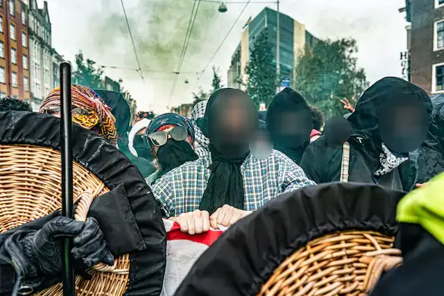 Een groep mensen met hoofddeksels en in sommige gevallen zonnebrillen op wordt midden op straat geconfronteerd door politieknuppels en -schilden. In de achtergrond stijgt een groene rookpluim op.