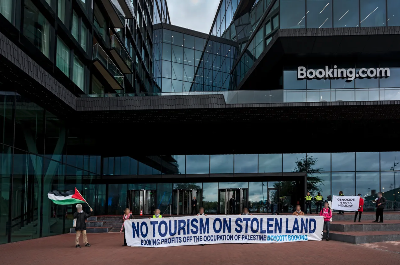 Een groep van ongeveer tien mensen staat voor een grotendeels glazen gebouw met op dat gebouw de tekst 'Booking.com'. De mensen houden Palestijnse vlaggen en twee spandoeken vast (beide in het Engels). Eén met de tekst: 'Geen toerisme op gestolen land. Booking profiteert van de bezetting van Palestina. Boycot Booking'. De ander met de tekst: 'Genocide is geen vakantieuitje'. Achter de mensen staan en stuk of vijf politieagenten en bewakers. De deuren bij de glazen ingang van het gebouwen hebben stuk voor stuk glasschade.