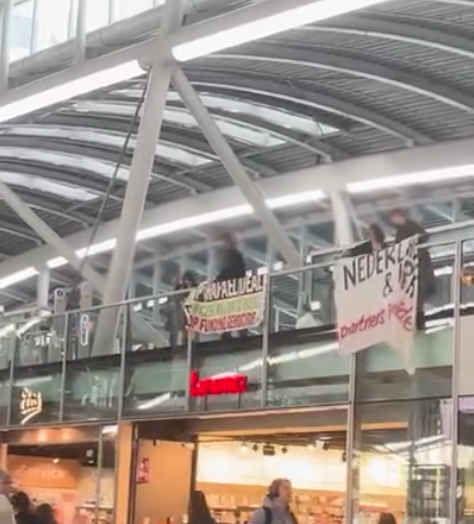 Een groep mensen staat op een ballustrade in een stationshal met een spandoek, en hgooit flyers naar beneden.