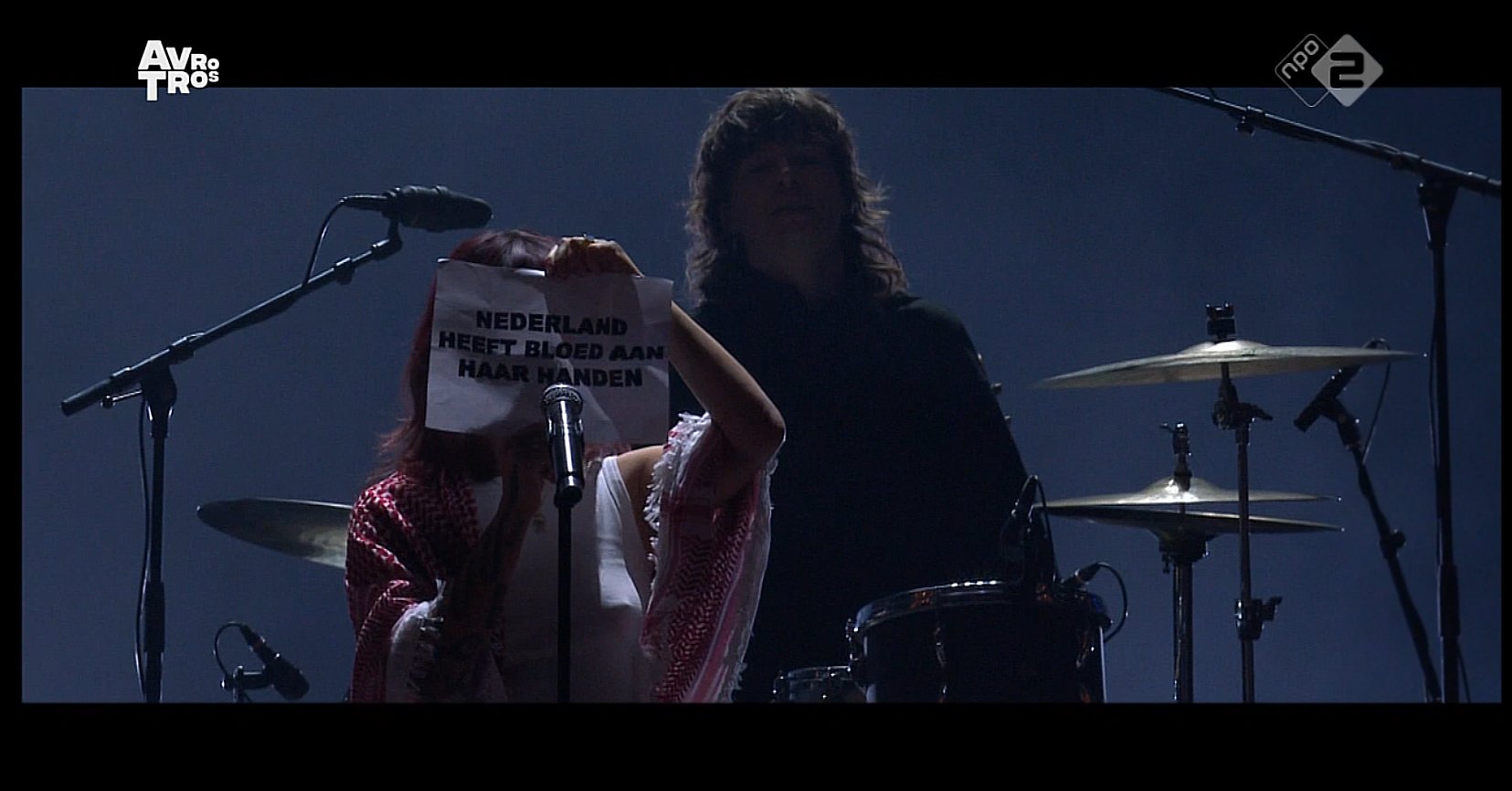 Een beeld van een concert, iemand achter een microfoon met een rood-witte keffiyeh, ze houdt een briefje op met daarop Nederland heeft bloed aan haar handen.