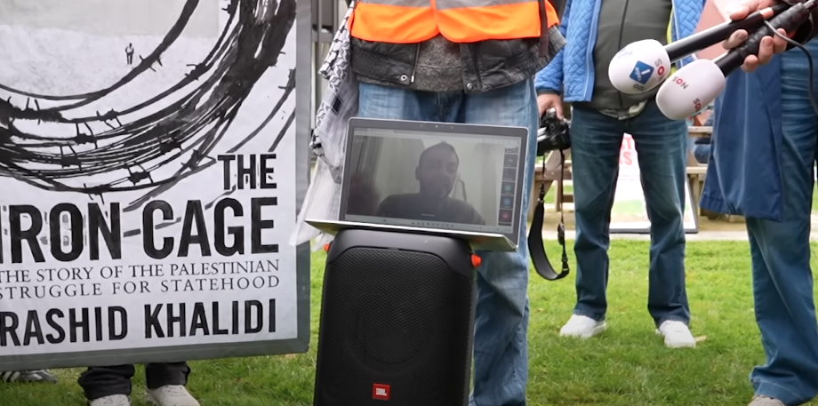 Een beeld met een laptop die balanceert op een verhoging, ergens buiten. Je ziet een stukje van een protestbord. Op de laptop is iemand te zien die via een video-verbinding aan het praten is.