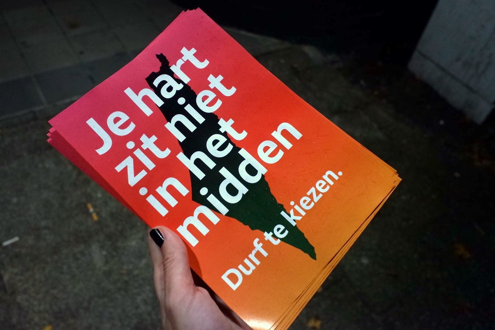 Een foto van iemand die een flyer ophoudt. Op de flyer staat: 'Je hart zit niet in het midden' en 'Durft te kiezen'. Op de achtergrond is de contour van Palestina te zien.